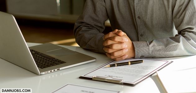A person sits at a desk with a closed laptop and a clipboard with a document and pen. Their hands are clasped, suggesting a formal setting.